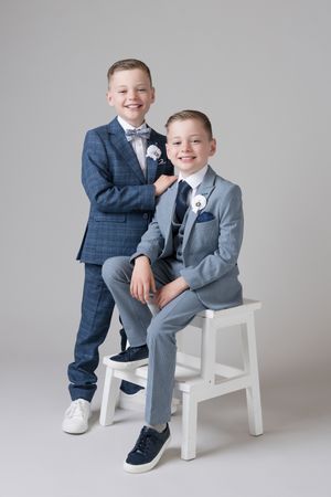 Two children in formal suits pose playfully on a white chair against a gray studio backdrop.