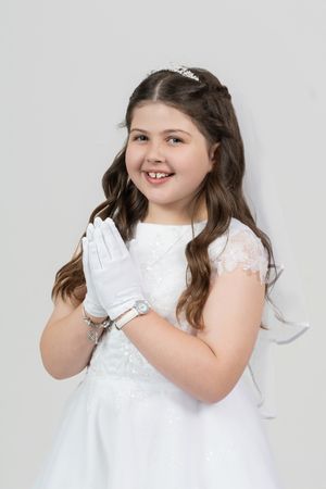 A child in a white dress and gloves poses sweetly against a light gray studio background.