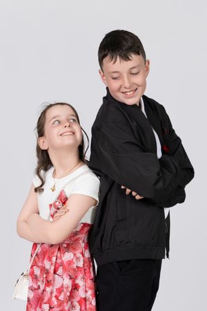 Two children stand back to back, one in a red floral dress and one in black jacket, both smiling and posing playfully.
