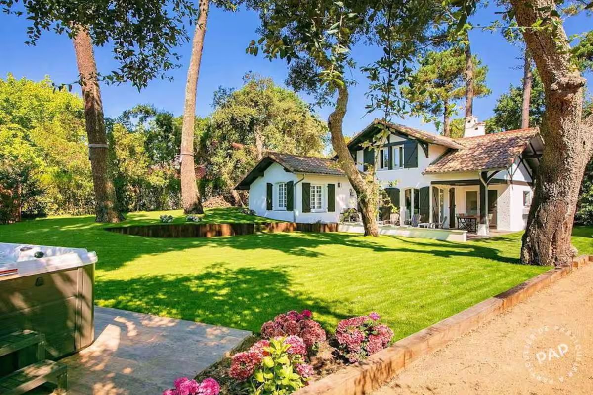 Modern beach house with pale blue exterior and manicured lawn surrounded by tall trees and pink flowers.