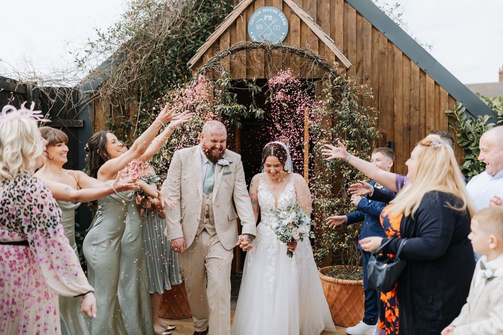 Wedding party celebrates with confetti toss outside rustic wooden barn with floral arch decorations.