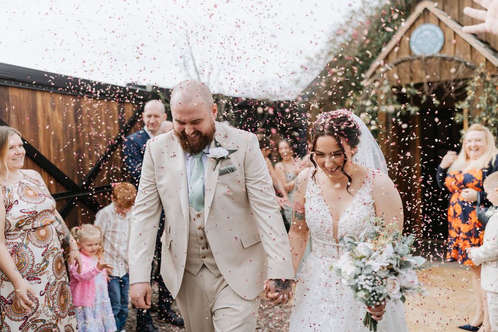 Wedding guests celebrate and throw confetti as a happy couple exits their ceremony at a rustic venue.