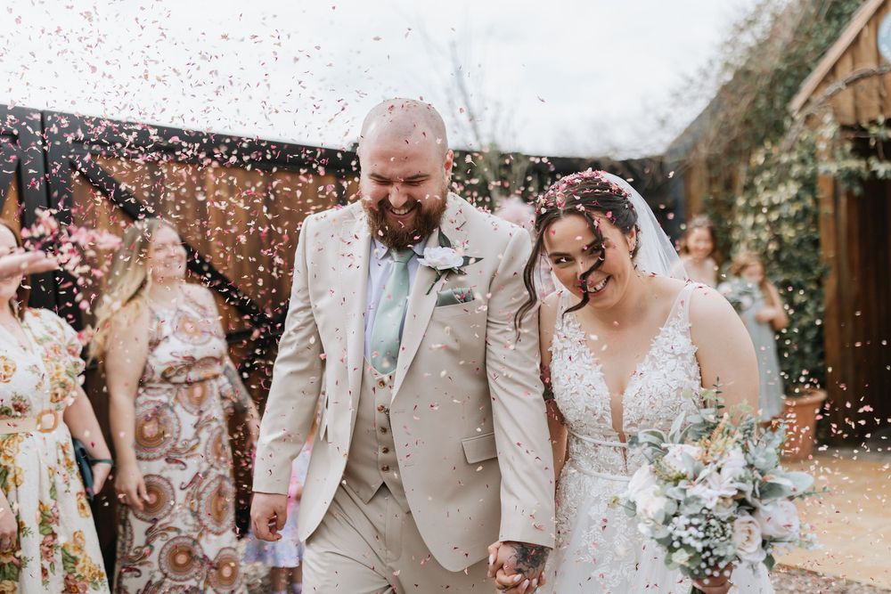 A newlywed couple walks through confetti shower during their outdoor wedding celebration at a rustic venue.
