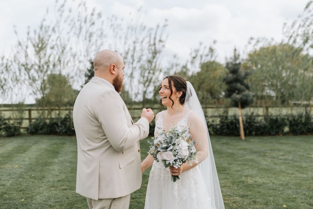Two people in wedding attire stand together on a green lawn holding a beautiful white floral bouquet.