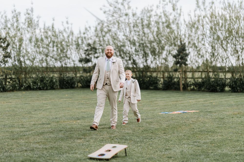 Two people in matching beige suits play cornhole on a green lawn with shrubs in the background.