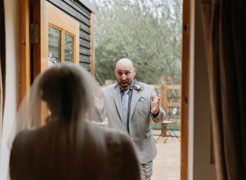 Person in a light gray suit gestures while having a conversation through a doorway with interior curtains visible.