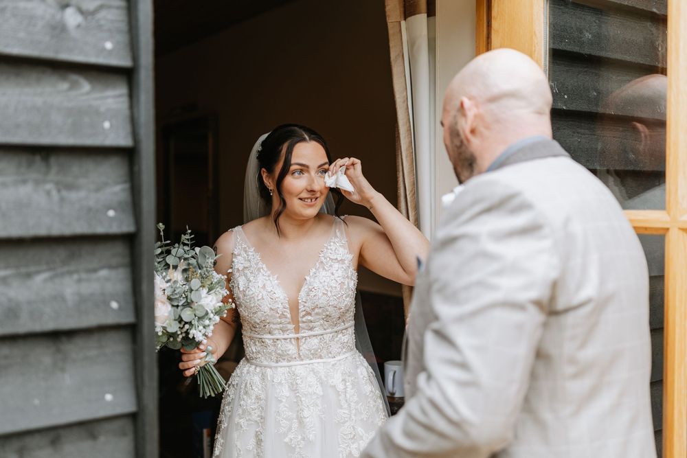 An emotional first look moment between a bride in a lace plunge wedding dress and a person in white at a rustic venue.