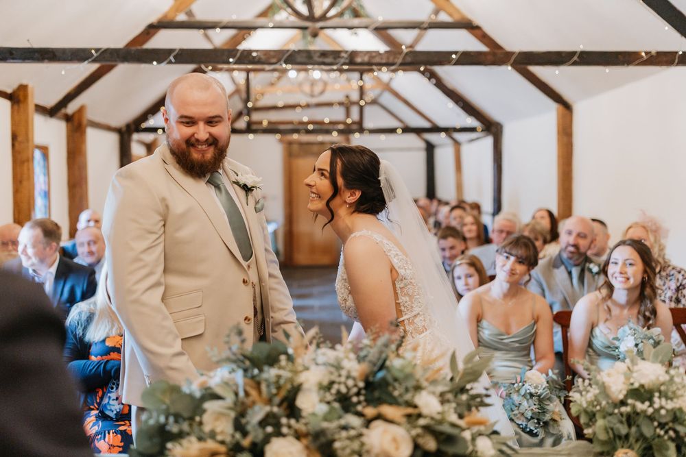 A wedding ceremony takes place in a rustic wooden barn venue with exposed beams, white walls and floral decorations.