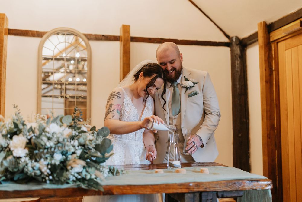 Couple performing unity candle ceremony with elegant floral decorations and wooden beams in rustic indoor setting.