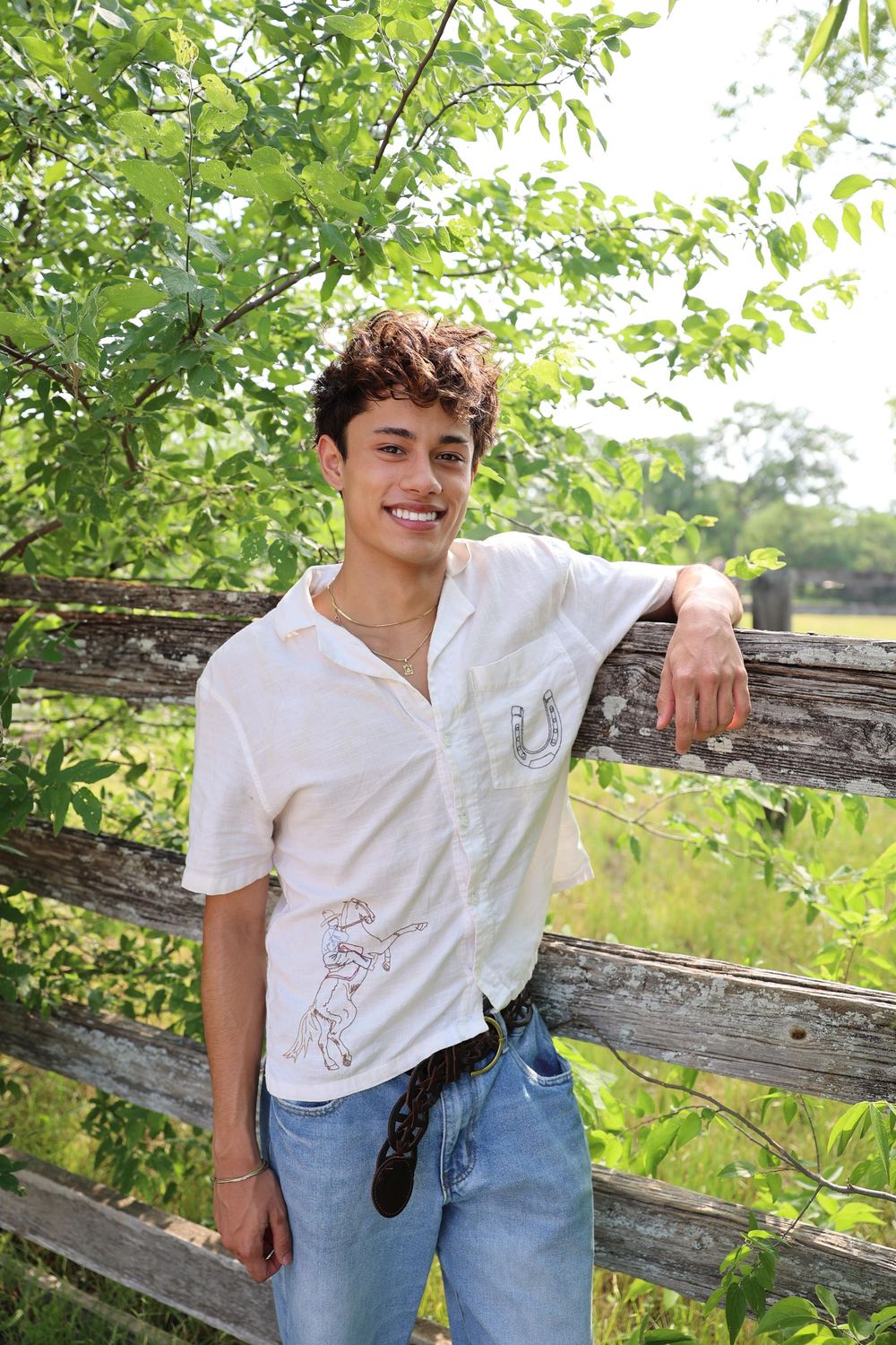 Person in white shirt and jeans leans casually against rustic wooden fence surrounded by green foliage.