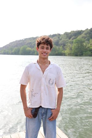 A person stands on a wooden dock wearing a white button-up shirt and jeans beside a scenic lake on a sunny day.