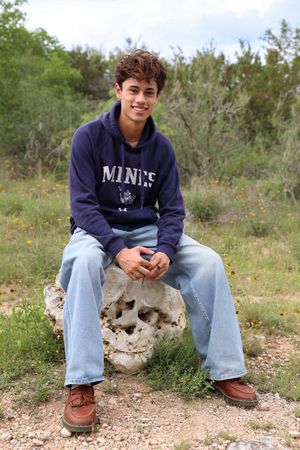 Someone wearing a Maine sweatshirt and jeans sits on a rock formation outdoors in a natural setting.