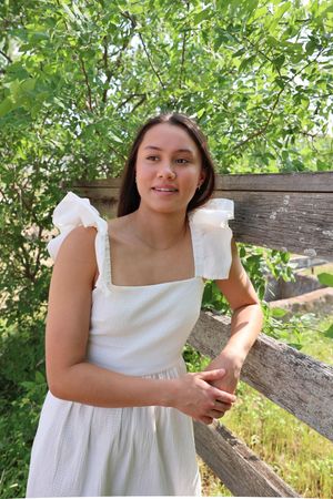 Someone wearing a white summer dress with ruffled sleeves poses next to a wooden fence in a sunny garden setting.