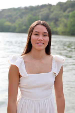 Portrait of someone in a white summer dress standing by a lake with green hills in the background.