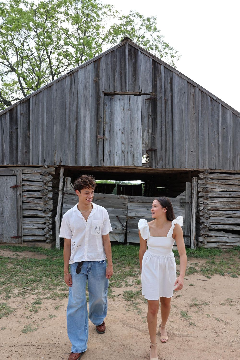 Two people stroll together in front of a weathered wooden barn with gray siding in a rustic outdoor setting.