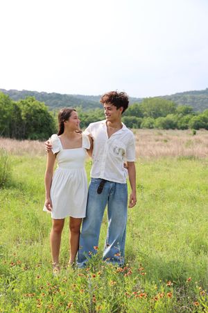 A couple dressed in summer clothes standing together in a sunny meadow filled with wildflowers.