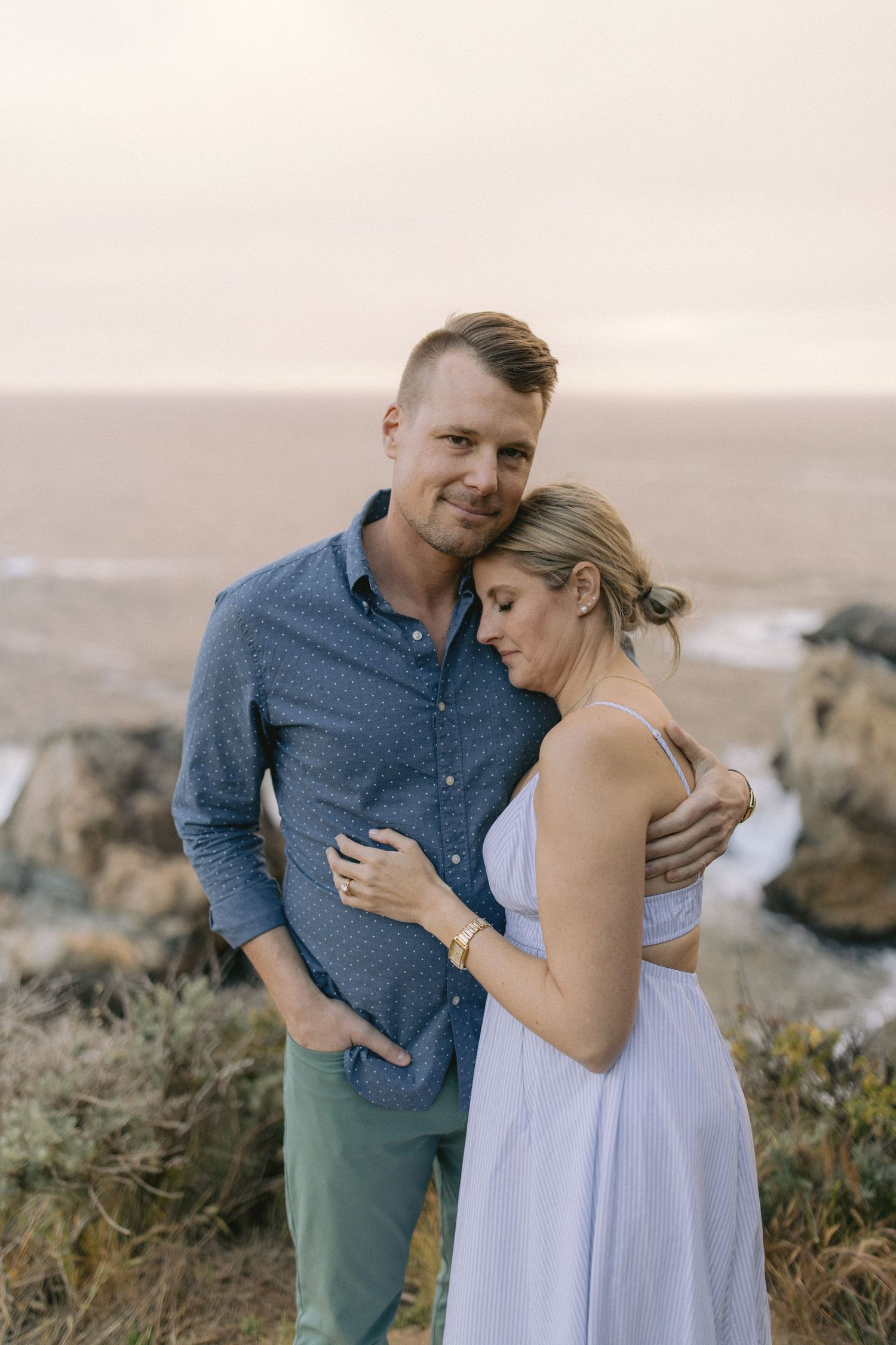 A romantic couple embraces on a coastal cliff at sunset, wearing blue and light colored clothing against a misty backdrop.