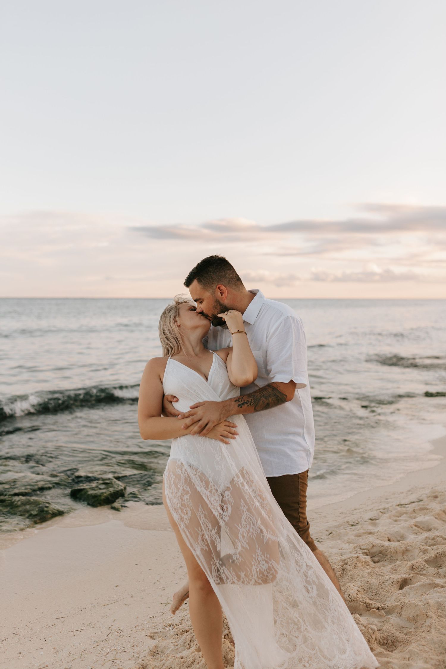 Couple sharing a romantic kiss on a sandy beach at sunset, woman in white lace dress, man in white shirt.