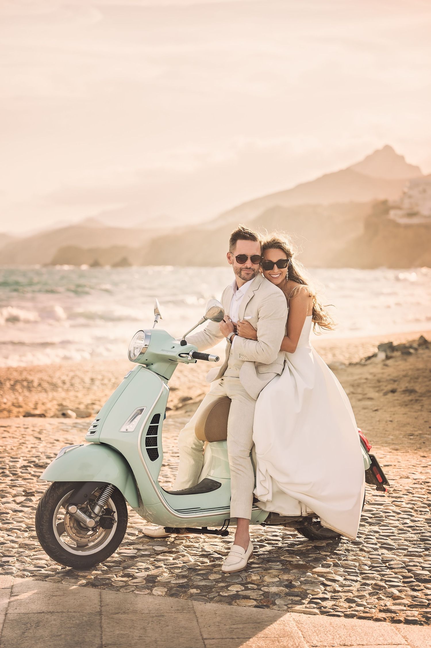 Newlyweds pose on mint green Vespa scooter during sunset at beach.