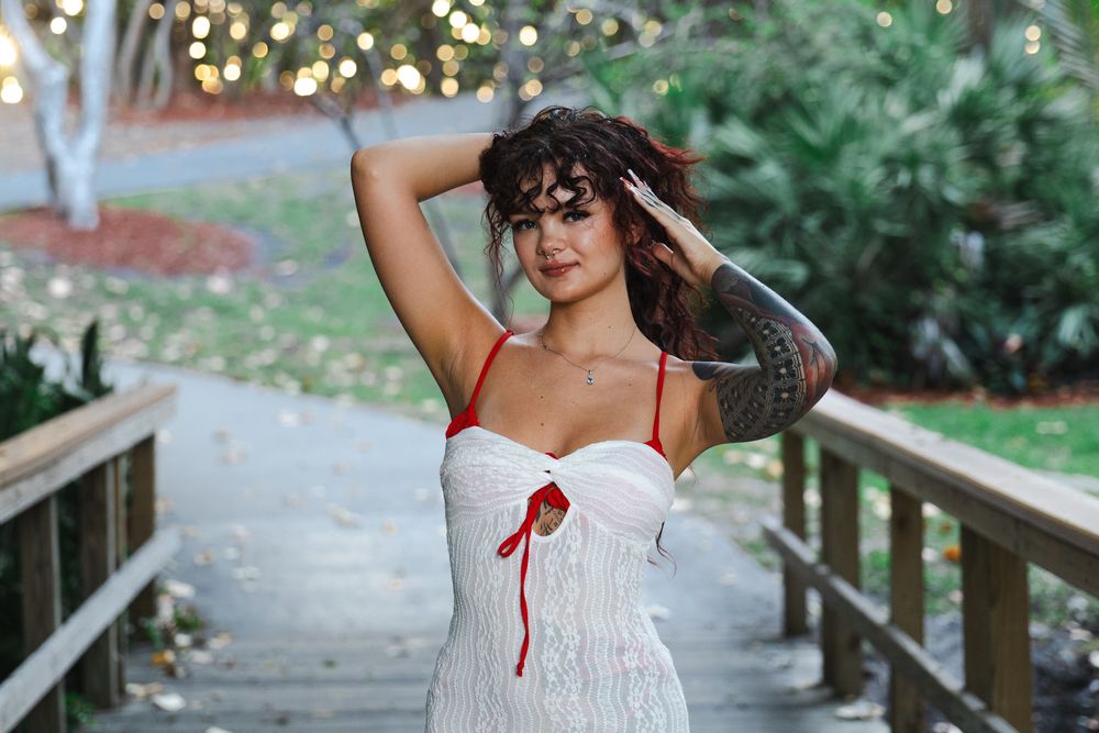 Figure in white summer dress stands on wooden walkway with garden backdrop.