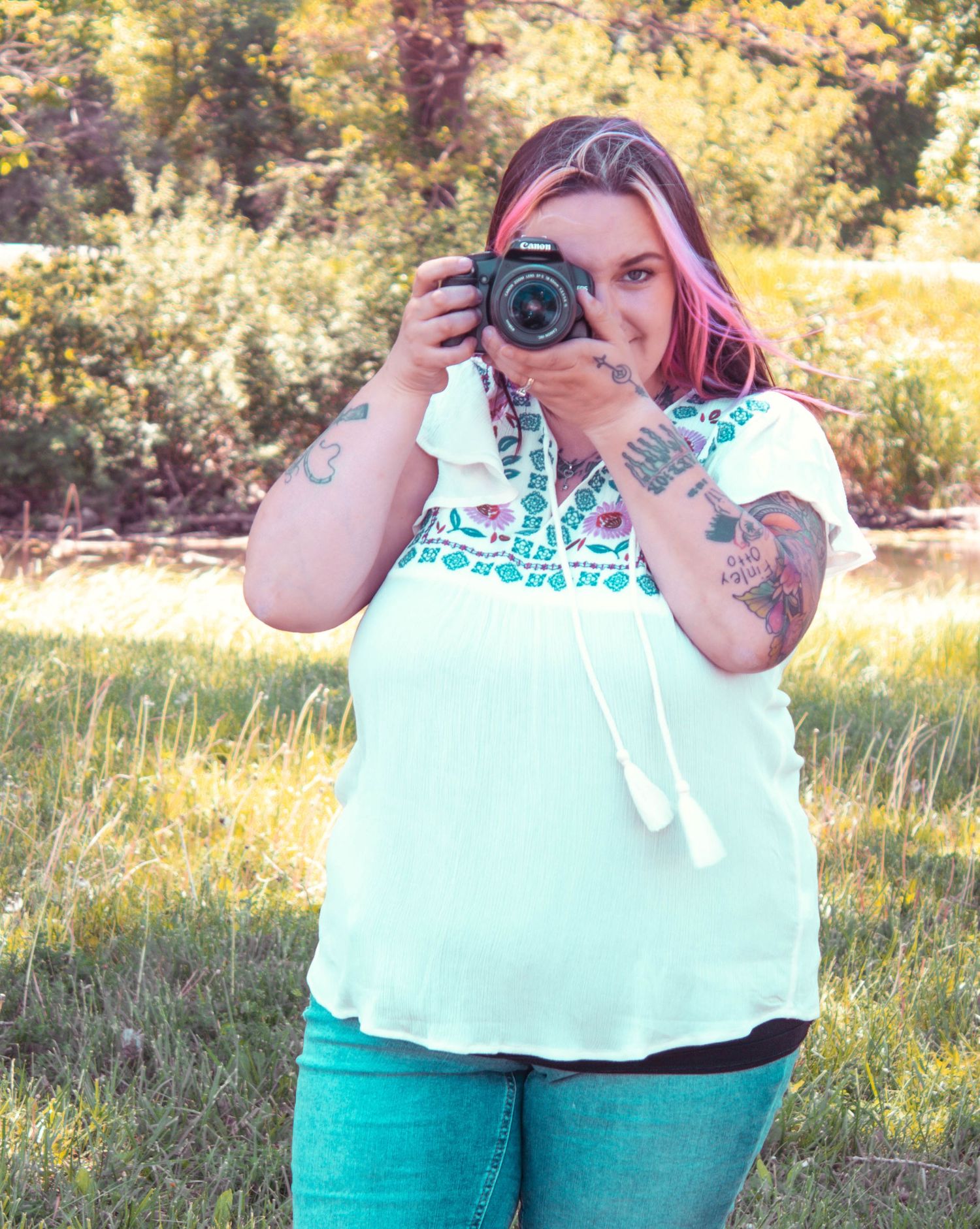 Photographer with tattoos takes picture with DSLR camera in sunny outdoor setting.