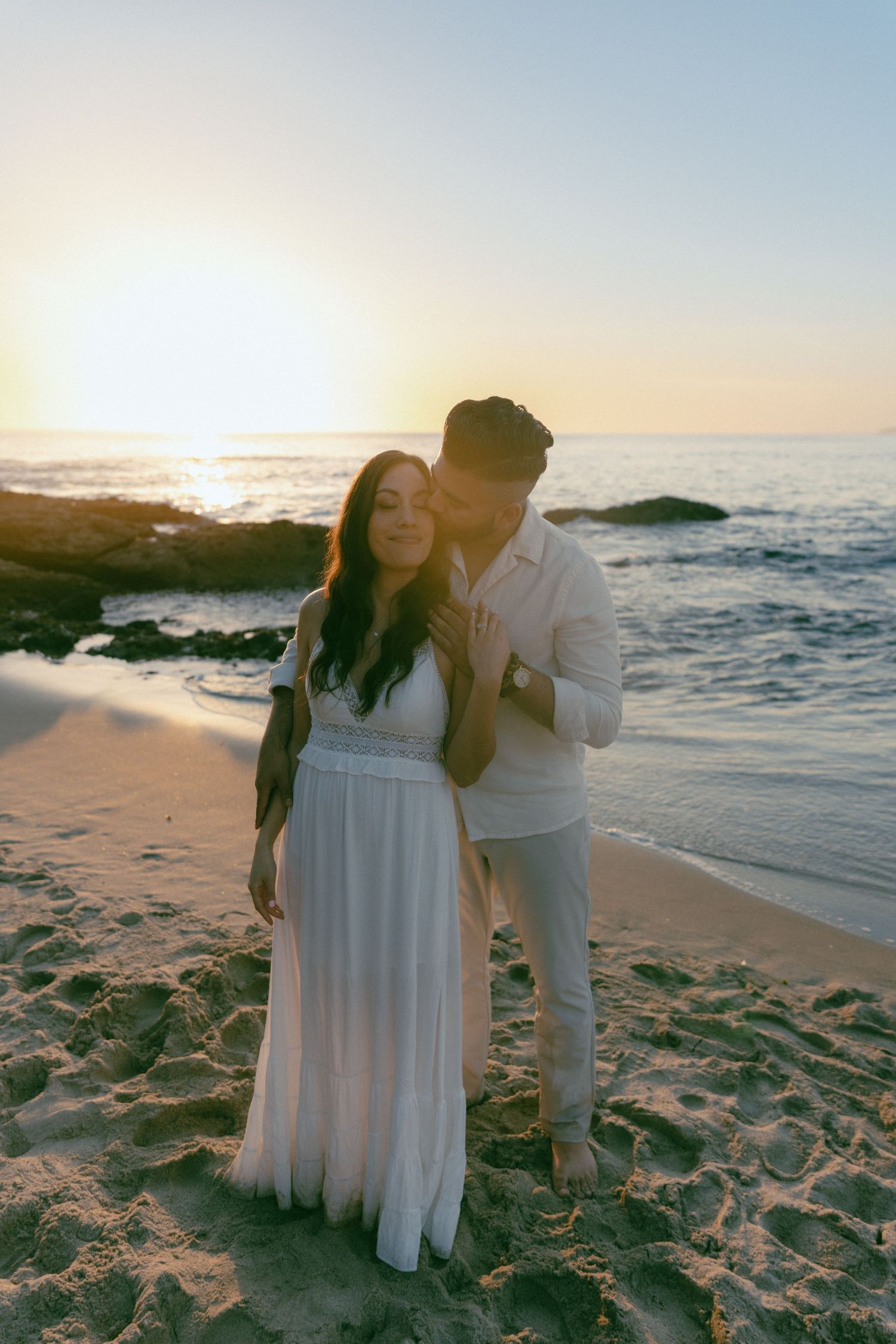 A romantic couple embraces on a sandy beach at sunset while waves crash on the rocky shore behind them.