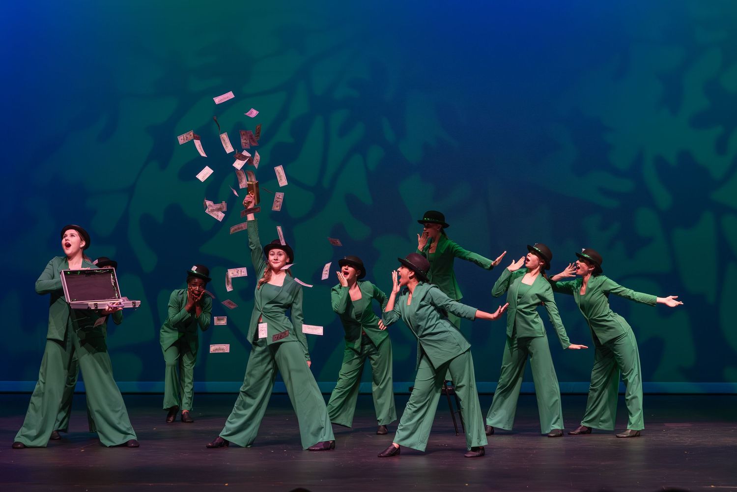 Dancers in flowing green costumes perform a synchronized routine on a dark stage with blue lighting effects.