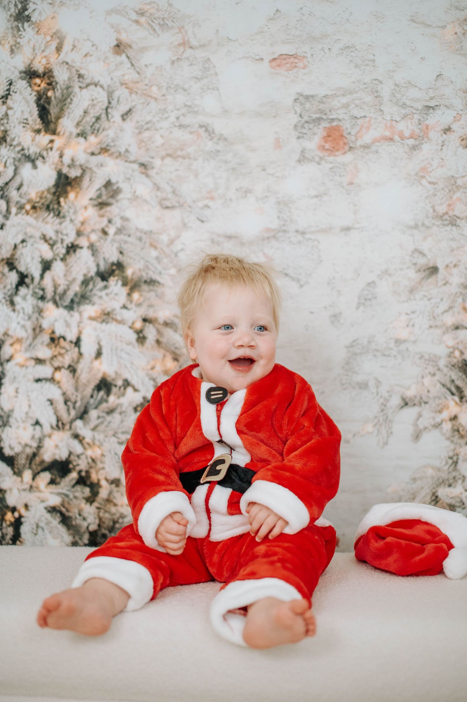 A baby in a red velvet Santa suit smiles and laughs while sitting on a white backdrop with winter decorations.
