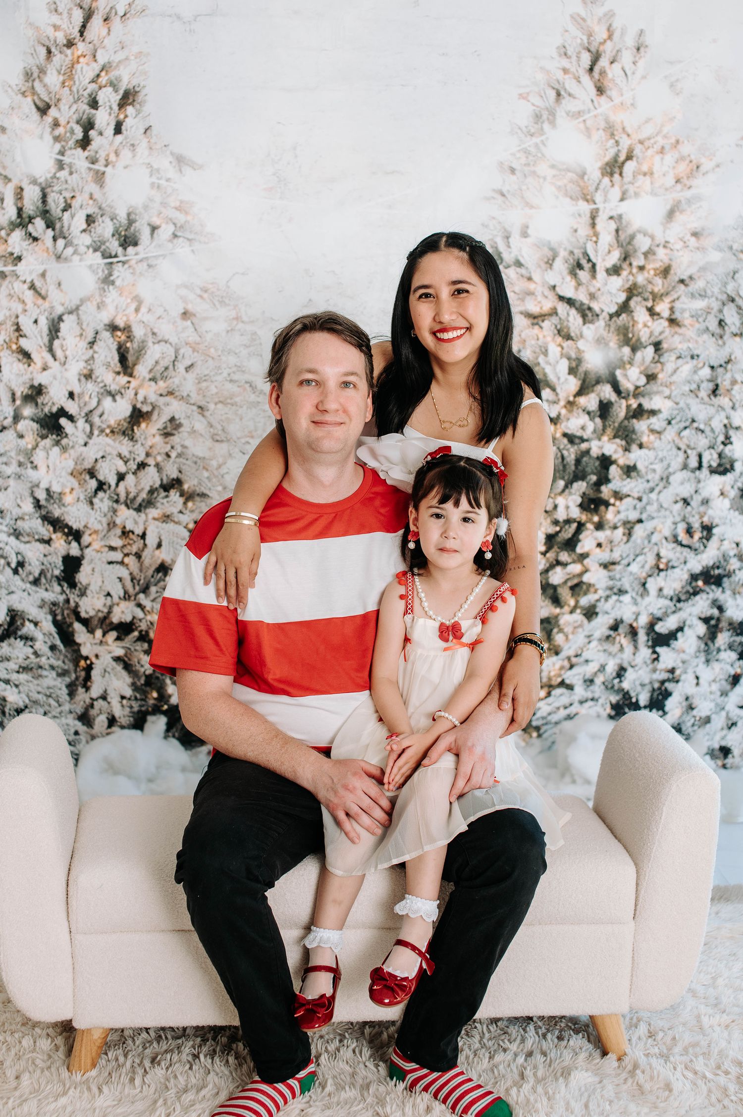 A family poses together on a cream couch against a backdrop of snowy white Christmas trees in a festive holiday setting.