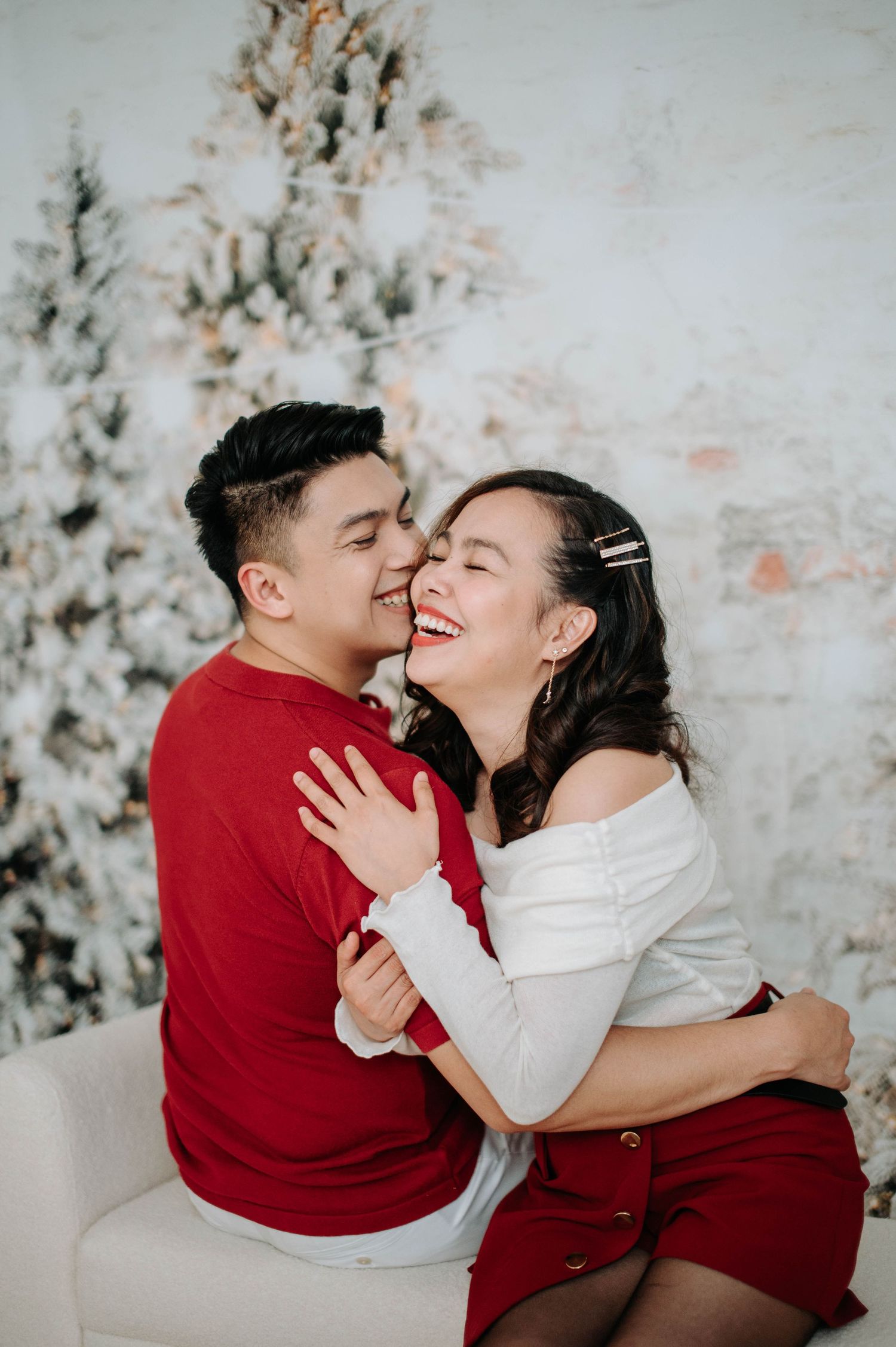 A couple in red and white outfits share a joyful moment together against a snowy winter backdrop with Christmas trees.