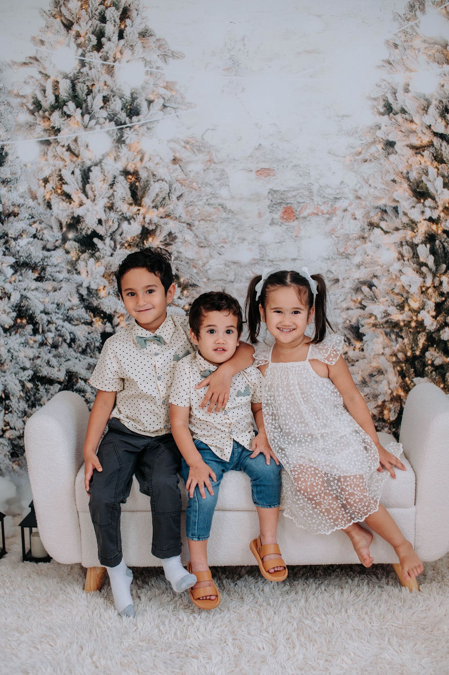 Three children sit together on a white couch against a backdrop of snowy winter trees and twinkling lights.