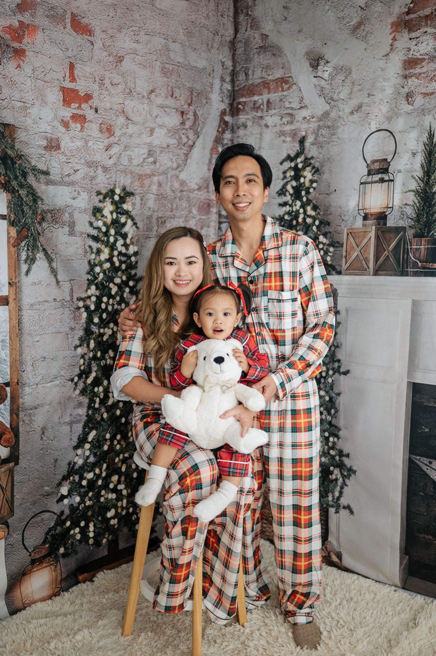 Family wearing matching plaid and checkered holiday pajamas pose together on white rug against rustic brick wall.
