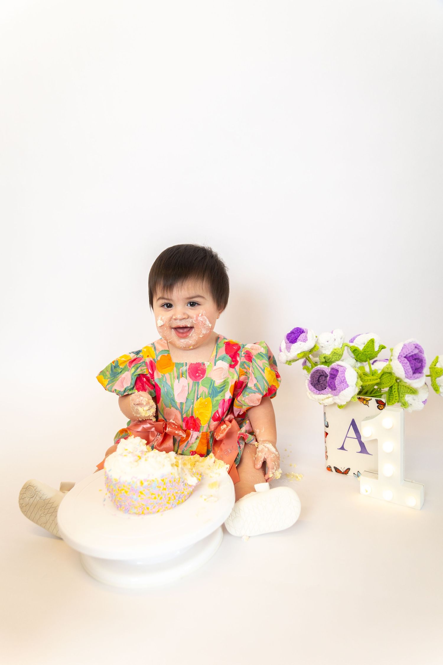 A smiling baby in a colorful floral dress sits next to a cake and purple flowers during a first birthday photoshoot.