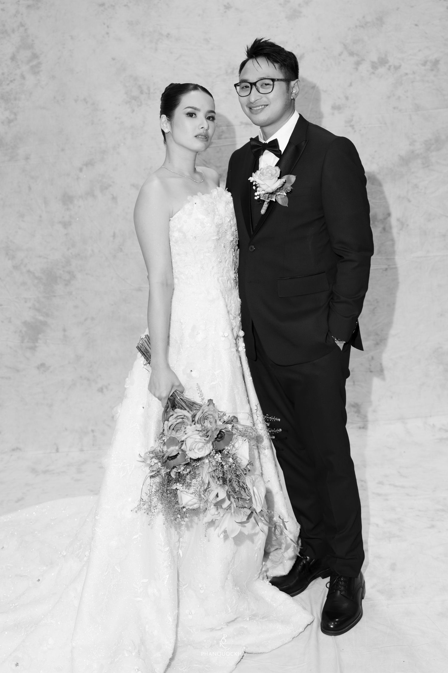 Black and white portrait of bride in strapless lace gown holding bouquet with groom in tuxedo and glasses.