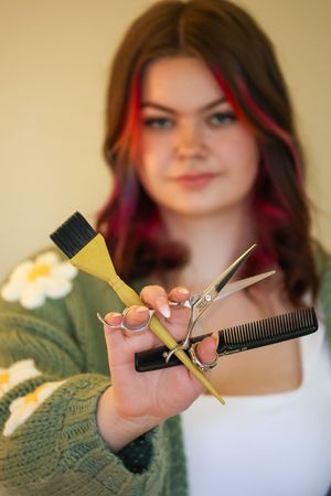 A hairstylist with red-streaked hair holds up professional styling tools including scissors and a dye brush.