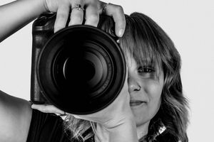A photographer peers through a large professional camera lens in black and white.