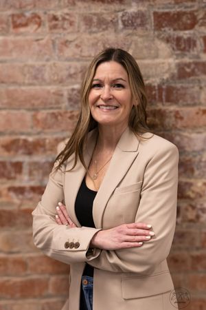 Professional headshots against a rustic brick wall background showing warm expressions in a beige blazer.