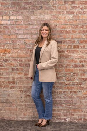 A person in a beige blazer and blue jeans poses against a rustic brick wall in three casual poses.