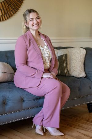 Professional wearing a pink blazer suit and floral top poses on a gray couch in a modern living room setting.