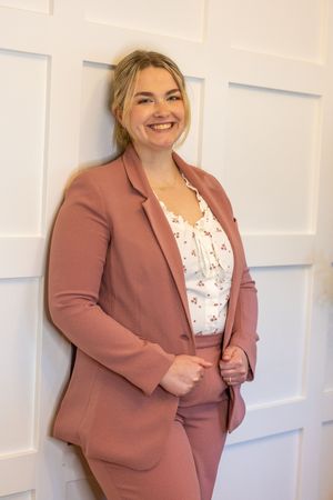 Professional wearing a mauve blazer and white floral top poses against white paneled wall.