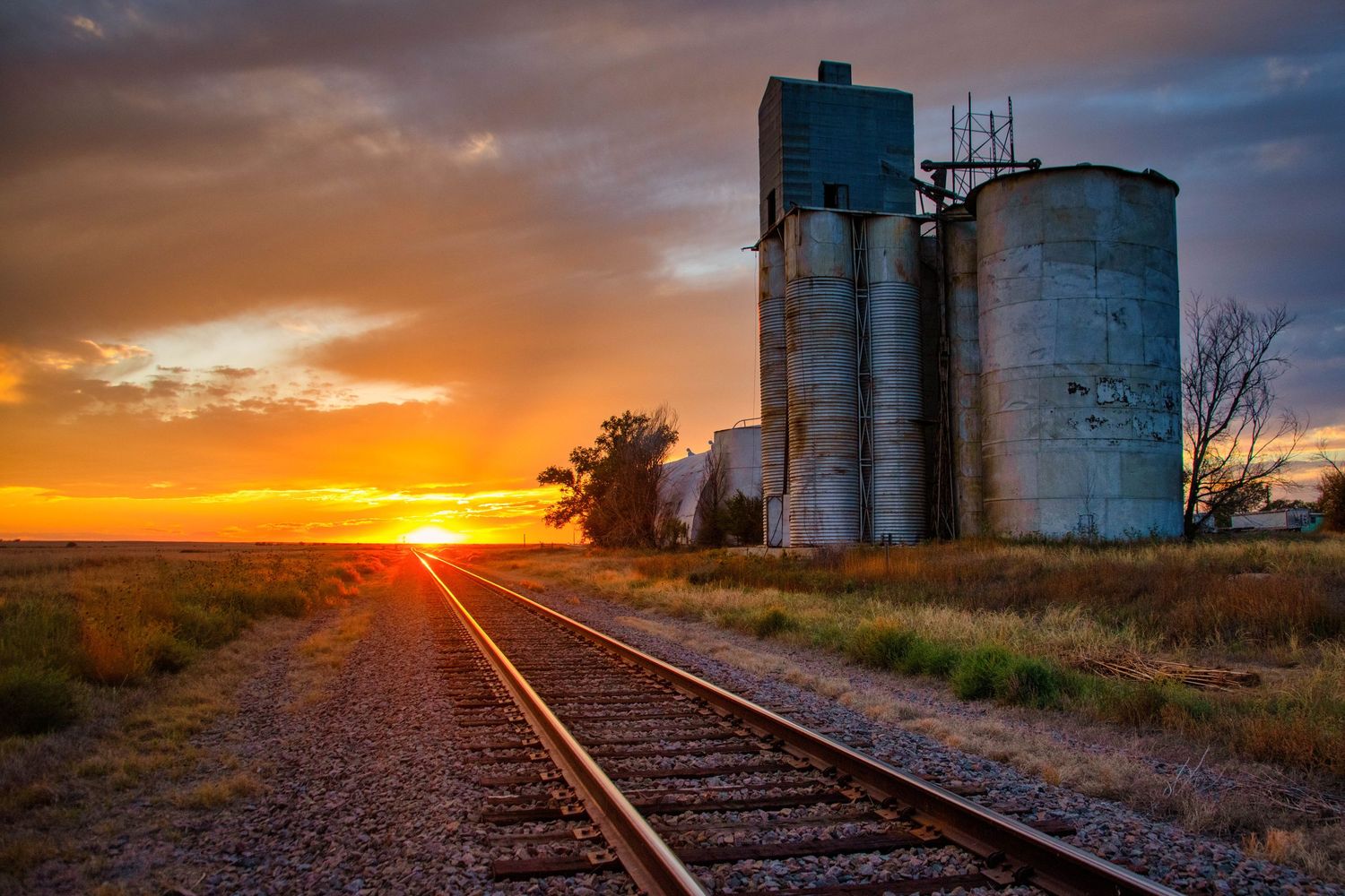 A dramatic sunset illuminates railroad tracks and grain silos in the rural countryside.