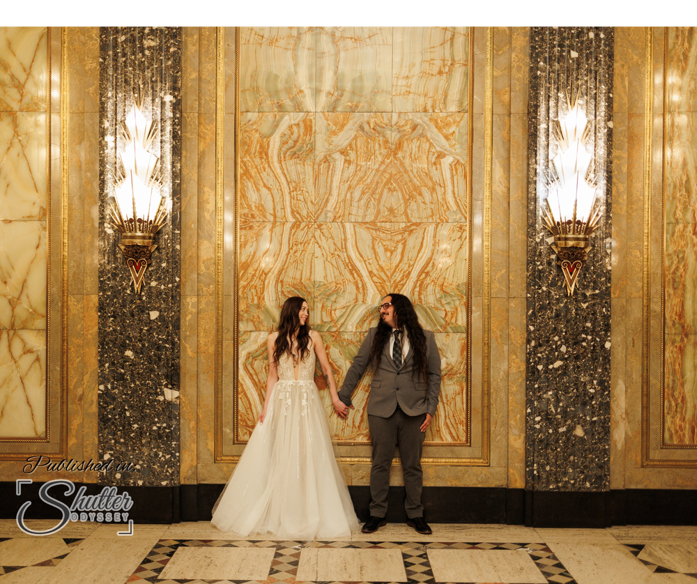 Newlywed couple holds hands while standing in an elegant art deco lobby with gold walls and marble columns.