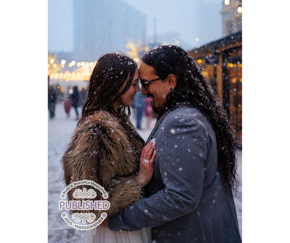 A couple shares an intimate moment in the falling snow outside a building with soft string lights in the background.