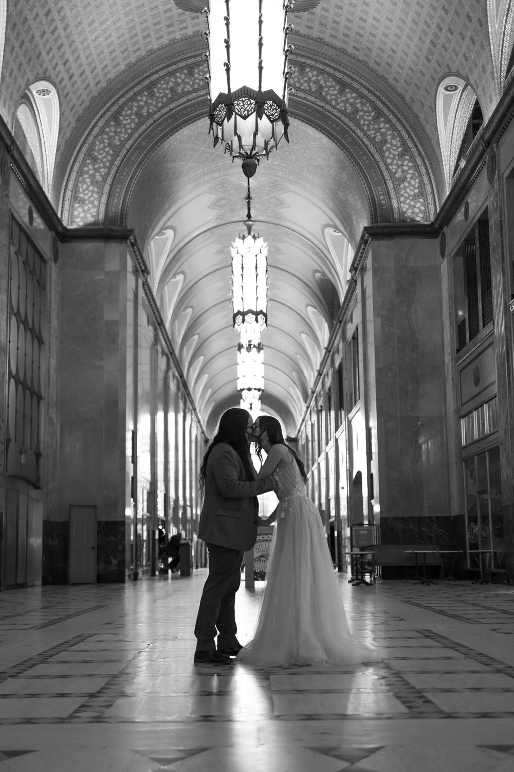 A romantic silhouette of a couple in formal attire standing in an elegant hallway with arched ceilings and ornate lighting.