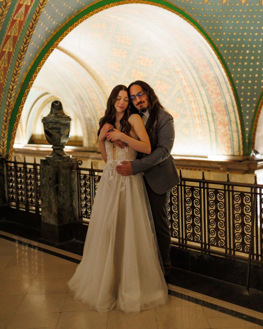 A couple in wedding attire embraces and poses for photos in an ornate arched hallway with decorative railings.
