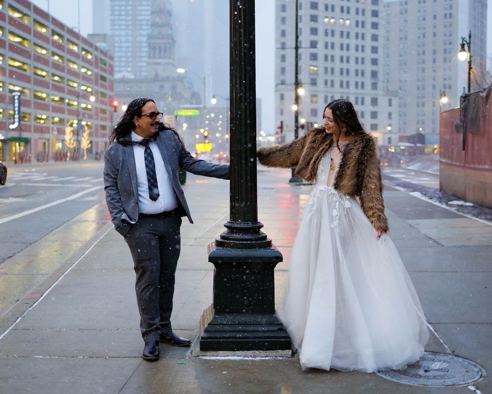 A couple poses together in a rainy urban setting with tall buildings in the background.