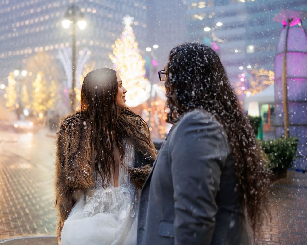 A couple walks together in the rain at night with city lights glowing in the background.