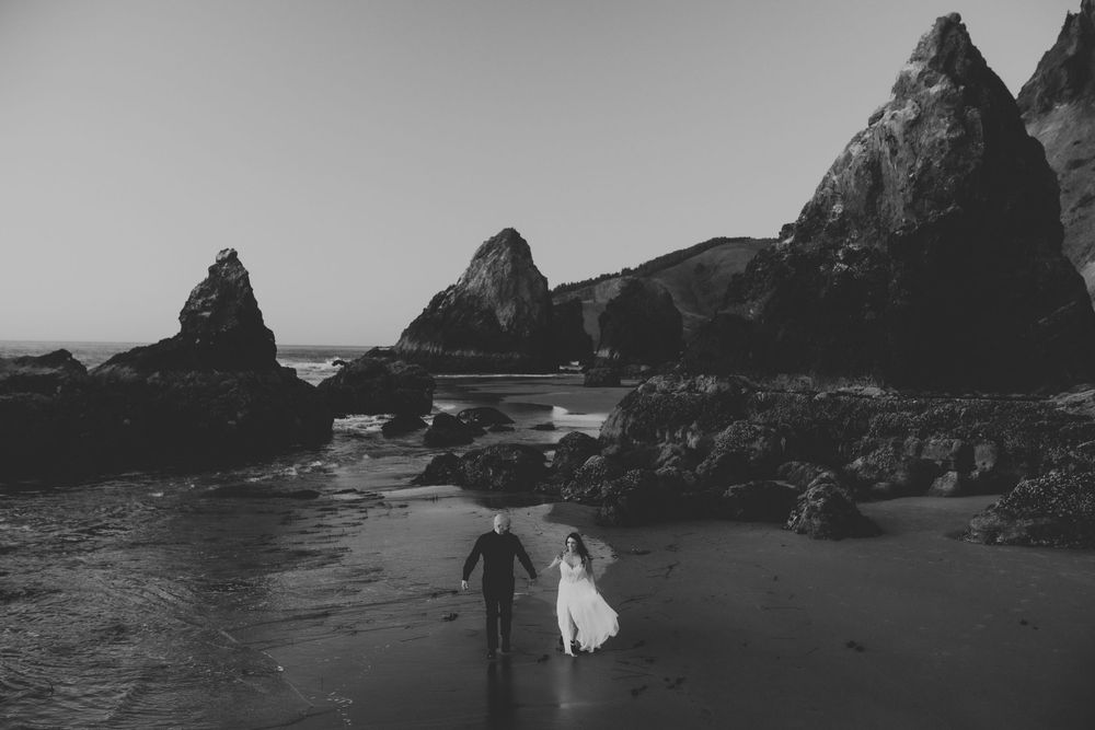 Dramatic black and white coastal landscape with rocky cliffs and sea stacks along a rugged beach at sunset.