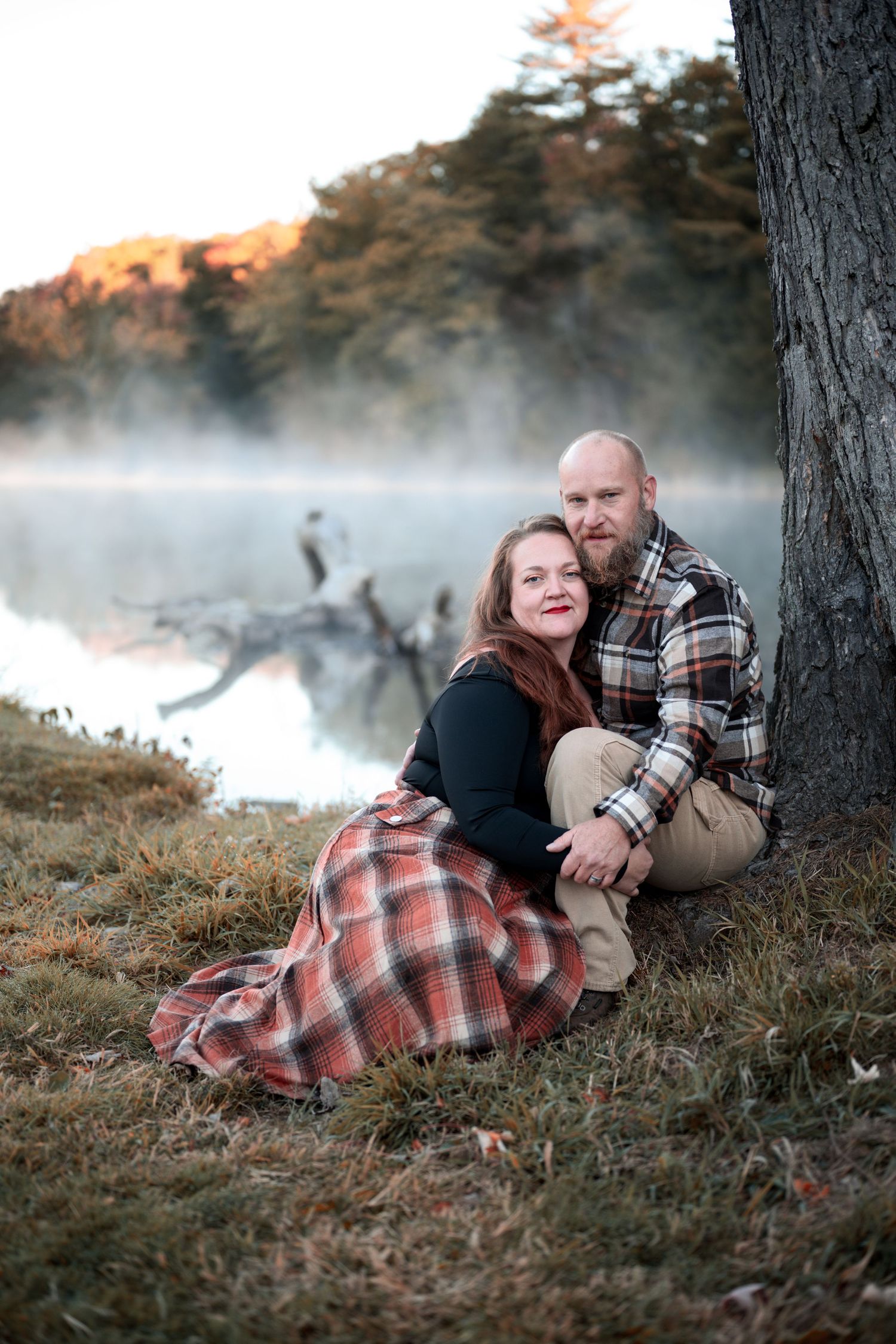Couple in plaid clothing sits by misty river at sunrise with fall trees and fog in background.