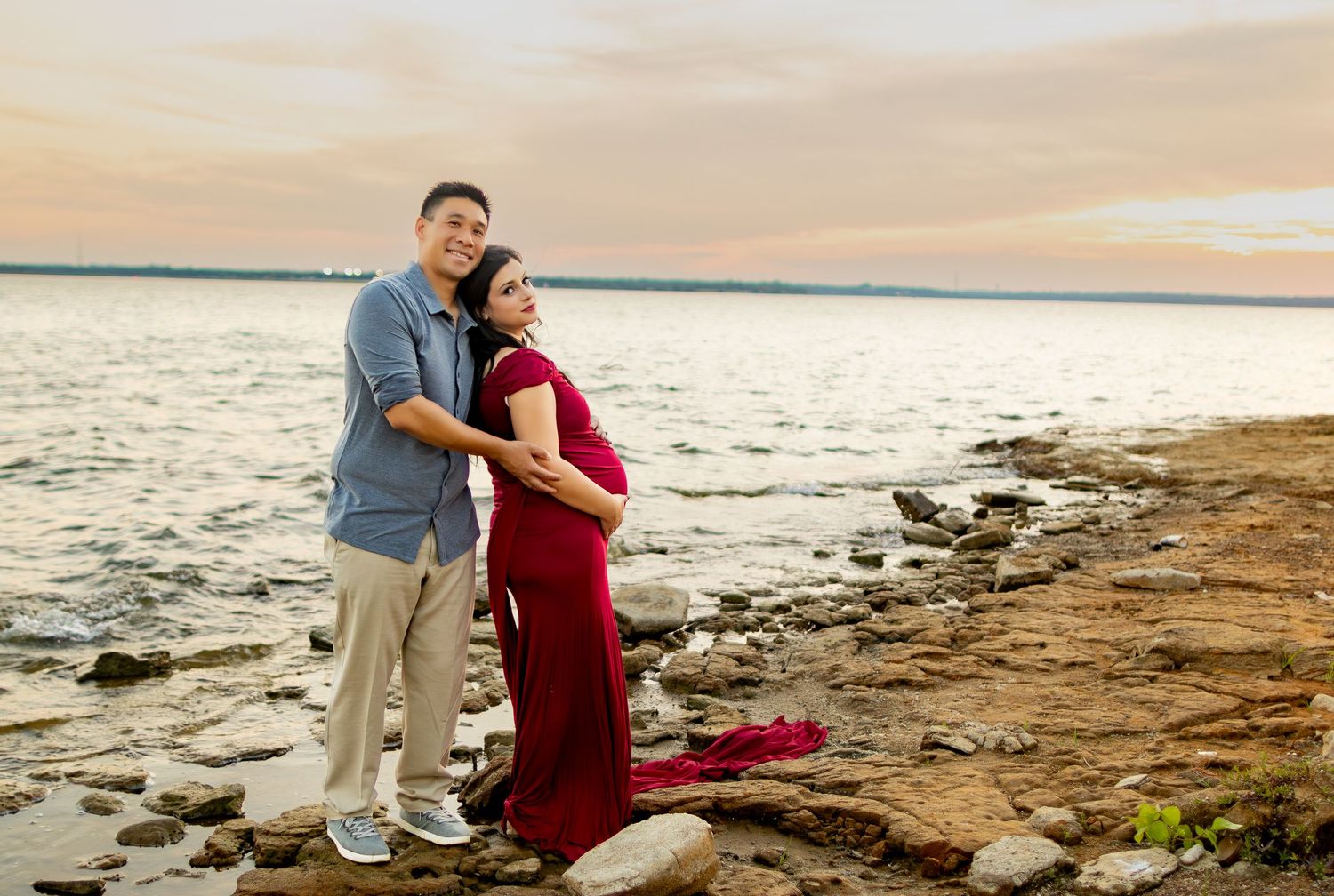Couple in casual attire embracing during sunset maternity shoot on rocky beach.